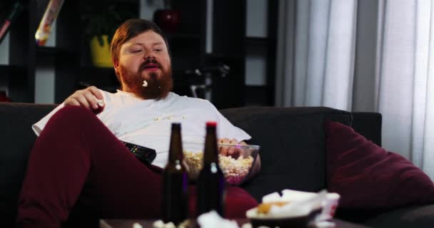 Homem gordo sorridente com barba assiste TV no quarto e come pipoca na mesa com cerveja. O gordo bebe cerveja. Conceito: Desnutrição, sedentarismo, descanso no sofá, obesidade — Vídeo de Stock