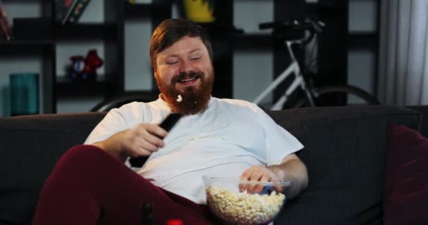 Homem gordo sorridente com barba assiste TV no quarto e come pipoca na mesa com cerveja. O gordo bebe cerveja. Conceito: Desnutrição, sedentarismo, descanso no sofá, obesidade — Vídeo de Stock