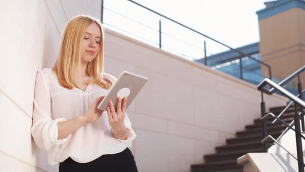 Mujer rubia bastante joven usando tableta dispositivo de computadora fuera en la calle. Mujer sonriendo, tocando en la pantalla táctil, viendo videos, noticias — Vídeo de stock