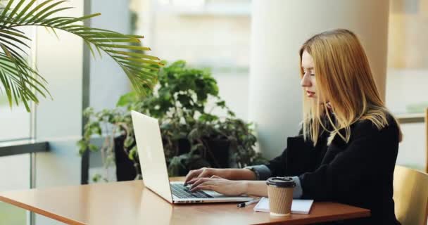 Fiatal szőke lány szerkezet-val egy laptop sittig Café asztalánál. Szabadúszó munkát keres, a nyomtatást, és kávét iszik — Stock videók