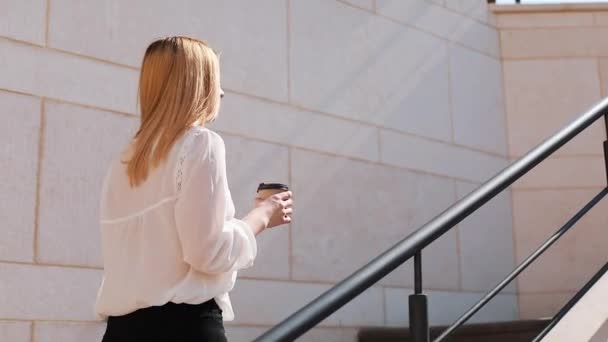 Joven mujer rubia de negocios camina arriba afuera en un día soleado cerca del edificio de oficinas — Vídeo de stock