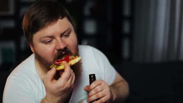 Gordo com barba come pizza sentada à mesa com cerveja e pipoca antes de um televisor — Vídeo de Stock