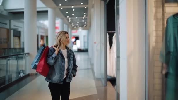 Mulher loira jovem e atenciosa caminha ao longo de uma janela de show com sacos no shopping — Vídeo de Stock