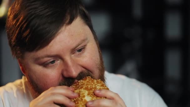 Gordo hombre mira la televisión y come una hamburguesa — Vídeo de stock
