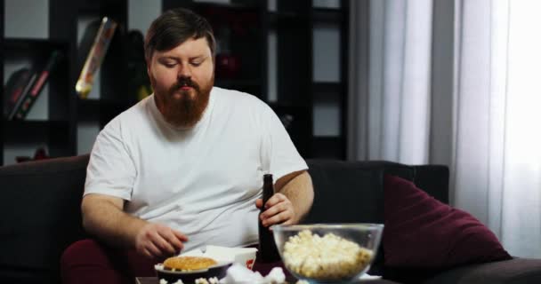 Um gordo engraçado a comer fast food, a beber cerveja e a treinar halteres. Ele está sentado em casa no sofá em frente à TV. Conceito de desnutrição, esporte, obesidade — Vídeo de Stock