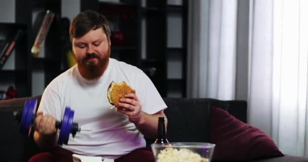 Um gordo engraçado a comer fast food, a beber cerveja e a treinar halteres. Ele está sentado em casa no sofá em frente à TV. Conceito de desnutrição, esporte, obesidade — Vídeo de Stock