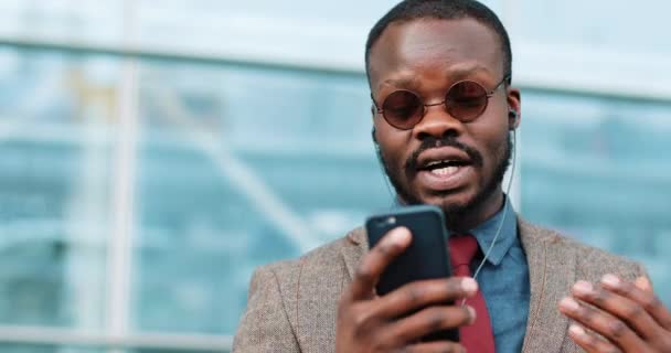 Retrato de jovem afro-americano falando usando a tecnologia de bate-papo por vídeo smartphone fica ao lado do centro de escritórios vestido com um terno de negócios com óculos de sol — Vídeo de Stock