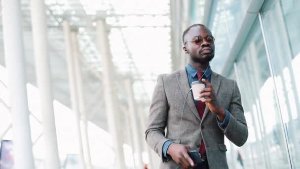 Happy african american businessman listening to music in headphones on smartphone, walking outside the office and funny dancing to the rythm. Drink coffee, funny dancing moves. Close up, slow motion — Stock Video