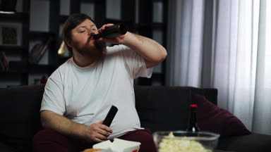 Şişman adam bira içiyor ve Tv izliyor. Yetersiz beslenme, bira alkolizm, obezite kavramı