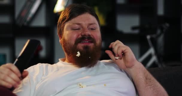 Hombre gordo sonriente con barba mira la televisión en la habitación y come maíz en la mesa con cerveza — Vídeo de stock