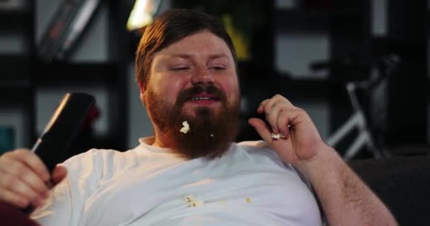 Souriant homme gras avec barbe regarde la télévision dans la chambre et mange pop-corn à la table avec de la bière — Video