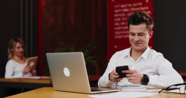 Jeune homme d'affaires souriant avec ordinateur portable et papiers appelant sur smartphone au bureau. Concept d'entreprise, de personnel, de communication et de technologie — Video