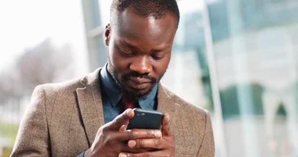 Attraktive afrikanisch-amerikanische Mann mit Smartphone in der Stadt. gutaussehender junger Geschäftsmann sendet per App SMS und lächelt glücklich. er steht in der Nähe von office cente auf der Straße — Stockvideo