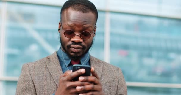 Привлекательный афроамериканец, использующий смартфон в городе. Красивый молодой бизнесмен слушает музыку через наушники, улыбаясь счастливо. Он стоит рядом с офисным центром на улице — стоковое видео
