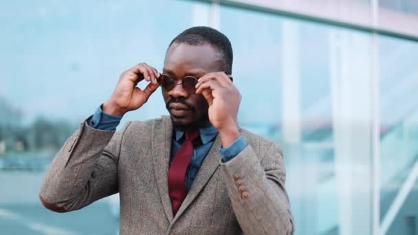 Homem de negócios negro africano em óculos de sol fica ao lado do centro de negócios - ao ar livre. O conceito de sucesso e negócios — Vídeo de Stock