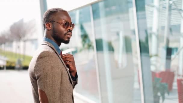 Beau Afro-Américain avec des lunettes de soleil homme d'affaires pose en costume moder dans la rue devant un bâtiment en verre. Le concept d'entreprise prospère — Video