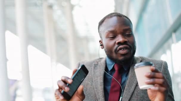 Empresário americano africano feliz ouvindo música em fones de ouvido no smartphone, andando fora do escritório e dançando engraçado para o ritmo. Bebe café, dança esquisita. Fecha, câmara lenta. — Vídeo de Stock