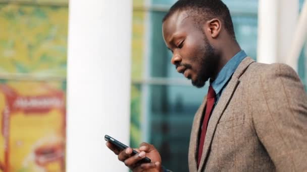 Glücklicher afrikanisch-amerikanischer Mann tippt eine Nachricht in sein Smartphone, als er auf der Straße in der Nähe eines Geschäftszentrums spaziert. Erfolgs-, Kommunikations- und Geschäftskonzept — Stockvideo