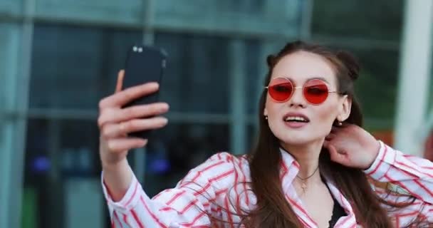 Bastante joven modelo en gafas de sol rojas toma una selfie en su teléfono inteligente — Vídeo de stock