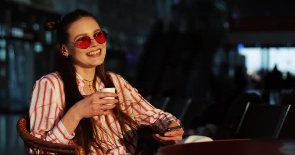 Encantadora joven en gafas de sol rojas bebe café en los rayos del sol de la noche — Vídeos de Stock