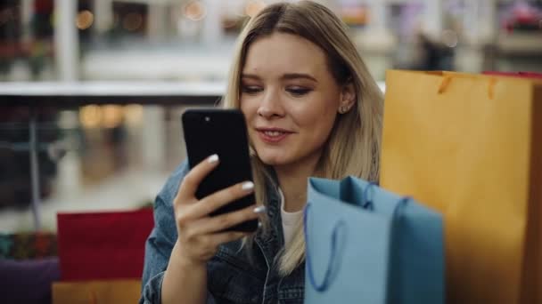 Гарненька блондинка друкує щось у своєму телефоні, сидячи в кафе з сумками — стокове відео