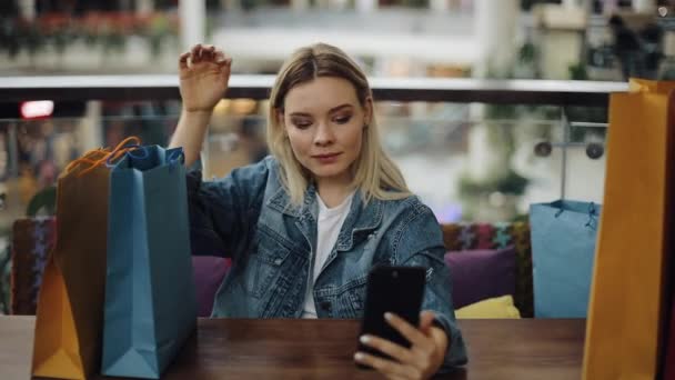 Hezká blondýnka dívka bere selfie na telefonu posezení v kavárně s nákupní tašky — Stock video