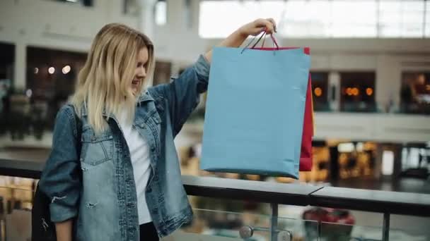 Menina loira feliz olha para seus sacos de compras coloridos em pé no shopping — Vídeo de Stock