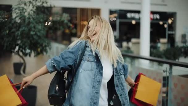 Charmig blond flicka i jeansjacka virvlar med shoppingkassar i gallerian — Stockvideo