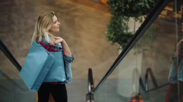 Joven mujer rubia con chaqueta vaquera sonríe bajando por las escaleras móviles en el centro comercial — Vídeos de Stock