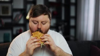 Şişman adam Tv izliyor ve evde kanepede oturan bir hamburger yiyor. Obezite, yetersiz beslenme kavramı