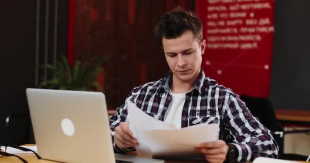 Feliz joven hombre de negocios leyendo papeleo en el escritorio en la oficina. Hombre casual sonriendo en el trabajo. Hombre trabajando en documentos. El concepto de contrato, negocio exitoso — Vídeos de Stock