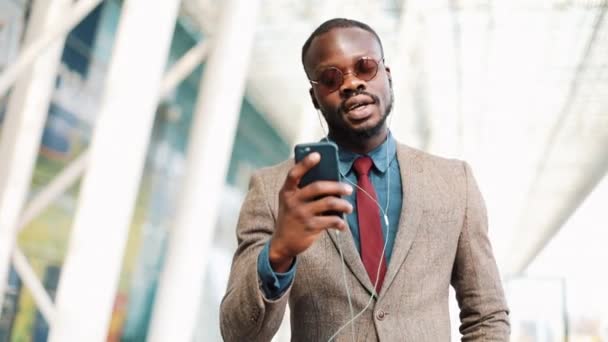 Hombre de negocios afroamericano con gafas de sol hablando por teléfono con auriculares cerca del edificio de oficinas. Negocios, personas, música, tecnología, ocio y estilo de vida — Vídeos de Stock
