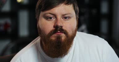 Okuma sakallı şişman adam doğruca kameraya benziyor. Dairesinde kanepede oturuyor