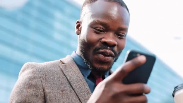 Счастливый афроамериканский успешный бизнесмен получает отличные новости на смартфоне. Он стоит рядом с офисным центром. Концепция приложения для смартфона — стоковое видео