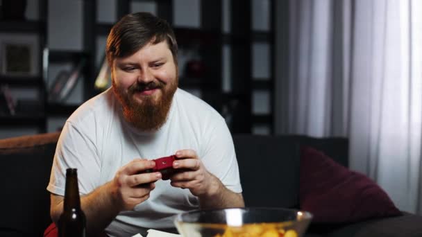 Толстяк играет на игровой консоли. Он сидит на диване в своей квартире, пьет пиво и ест фаст-фуд. Концепция видеоигр и недоедания, ожирения — стоковое видео