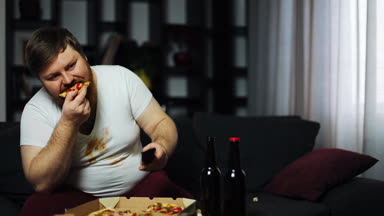 Pizza yiyip kanepede oturan bira içmeyi zayıf iradeli kilolu adam. Yetersiz beslenme, spor, obezite kavramı