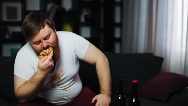 Слабый толстяк ест пиццу и пьет пиво, сидя на диване. Концепция недоедания, спорта, ожирения — стоковое видео