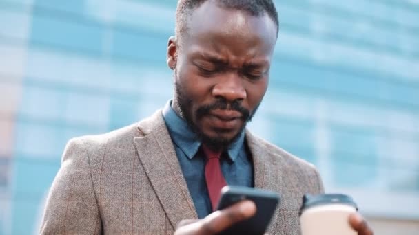 Уставший и напряженный афроамериканский бизнесмен читает что-то в своем смартфоне, стоящем снаружи. Тексты смс с использованием приложения на смартфоне в городе . — стоковое видео