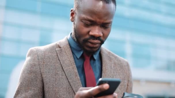 Stanco e stressato uomo d'affari afroamericano legge qualcosa nel suo smartphone in piedi fuori. Uomo sms sms utilizzando app su smartphone in città . — Video Stock