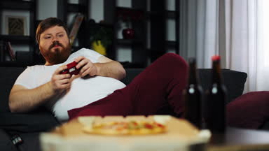 Şişman adam konsolda oyun oynuyor. O kanepede onun daire içecekler bira oturuyor ve hızlı yemek yiyor. Video oyunları ve yetersiz beslenme, obezite kavramı