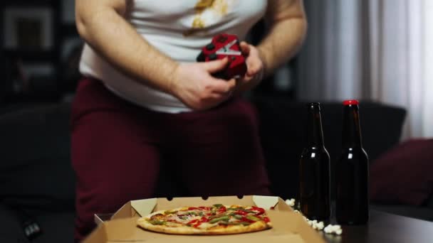 Der dicke Mann spielt an der Spielkonsole. Er sitzt auf der Couch in seiner Wohnung, trinkt Bier und isst Fast Food. Konzept der Videospiele und Mangelernährung, Fettleibigkeit — Stockvideo