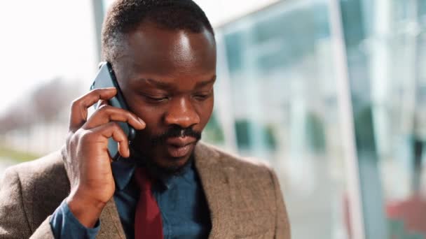 Hombre de negocios afroamericano habla con alguien en su teléfono inteligente de pie en la calle cerca del centro de oficinas. Negocios, personas, comunicación, tecnología, ocio y estilo de vida . — Vídeo de stock