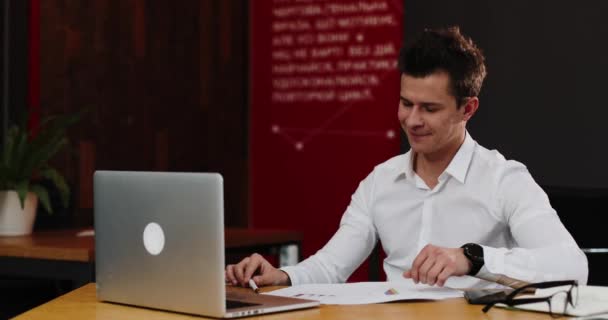 Ο άνθρωπος μιλά για την βιντεοκλήση στο λάπτοπ του στο γραφείο. Νέος ελκυστικός επιχειρηματίας σε ένα λευκό πουκάμισο που δείχνει σκατά o μετά την κλήση βίντεο — Αρχείο Βίντεο
