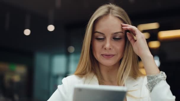 Mujer sonriente se arregla el pelo sentada en un centro comercial y leyendo una tableta — Vídeos de Stock