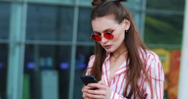 Genç kadın bir alışveriş merkezi yakınındaki telefon ayakta mesaj yazarak. Kadın pembe güneş gözlüğü güneşli gün bloglama Internet sörf sanal dünya mobil cihaz yaz tatili