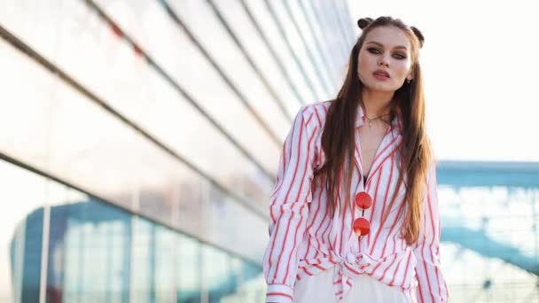 Impresionante modelo de moda con peinado y gafas de sol rojas frente a una pared de vidrio y posa para la cámara — Vídeo de stock