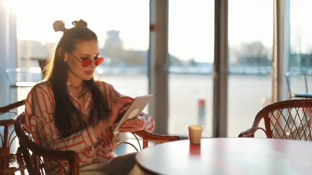 Mujer atractiva joven en gafas de sol rojas con tableta sentada en la cafetería. Hermosa chica en el aeropuerto o centro comercial con la tableta — Vídeos de Stock