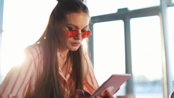 Mujer atractiva joven en gafas de sol rojas usando tableta con pantalla táctil en un café. Hermosa chica en el aeropuerto o centro comercial con la tableta — Vídeos de Stock