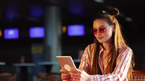 Young attractive woman in red sunglasses with tablet computer sitting in cafe. Beautiful girl in airport or shoppping mall with tablet — Stock Video