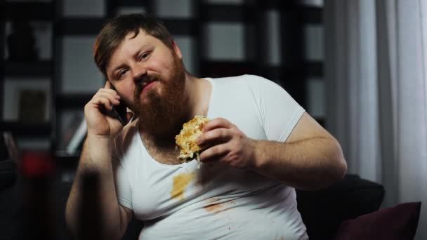汚れた脂肪ひげを生やした人間が話をスマート フォン、ハンバーガーを食べて、ソファに座ってビールを飲んでします。栄養失調、食糧配達、通信、肥満の概念 — ストック動画